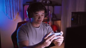 A cheerful young asian man with headphones using a smartphone in a neon-lit gaming room at night.