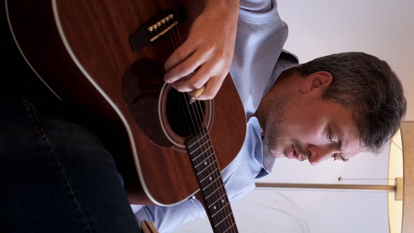 Vertical video. Composing song. Musician creativity. Concentrated man sitting writing in journal playing acoustic guitar new melody chords in light home interior.