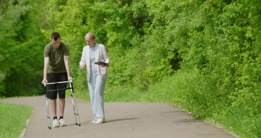 A woman assists a young man as he walks with a walker, undergoing rehabilitation after an injury.