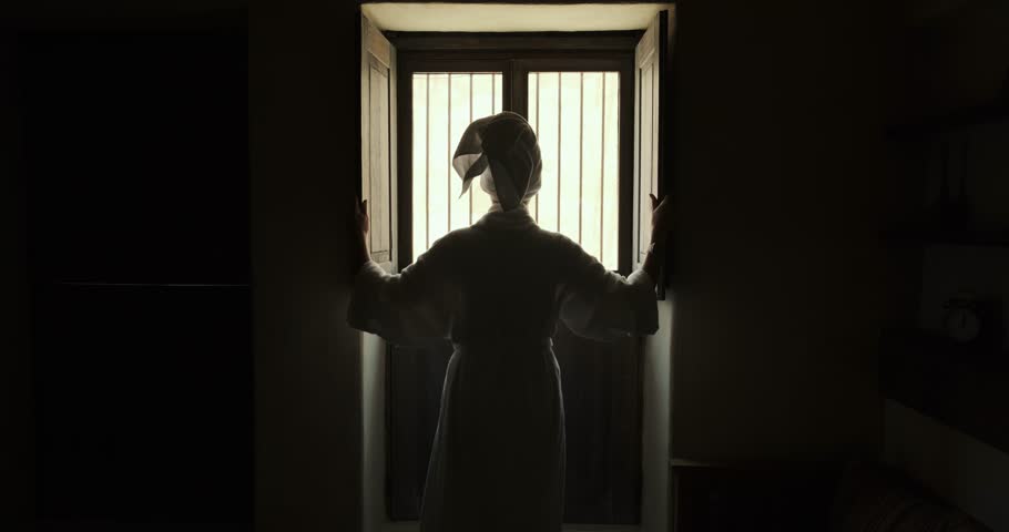 Woman in robe room of an old hotel looks out window and closes it. young woman stands in room by window and closes shutters. Girl close curtains. Young female approaches window and closes curtains