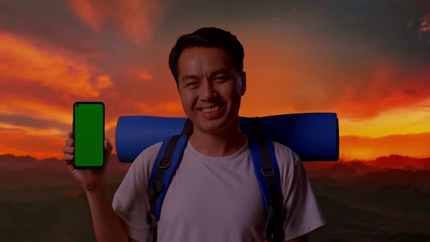 Close Up Of Asian Male Hiker With Mountaineering Backpack Smiling And Showing Mock Up Green Screen Smartphone While Standing On Top Mountain During Sunset Time