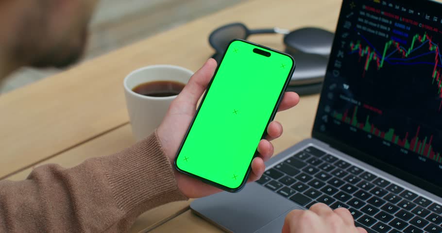 The man uses a mobile phone with green screen. He uses the laptop sitting at his desk. Graphs on the screen. An unrecognizable person