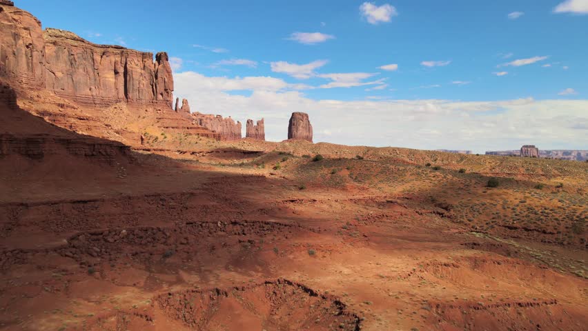 Experience an aerial perspective of the Monument Valley Desert with towering mountains in the background. The vast expanse of arid land stretches into the distance under the clear blue sky.
