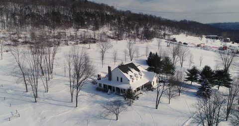 A high angle forward flyover of a typical snow-covered farmhouse in rural Pennsylvania in the winter.  	
