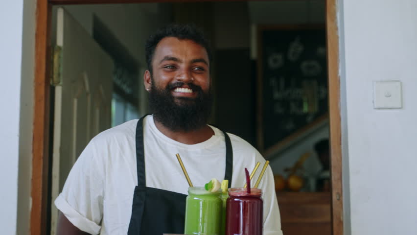 Black smiling waiter carries healthy fruit smoothies and putting drinks on the table in cafe on Bali. Cheerful waiter serving customers in the tropical restaurant. Waiter delivering cocktails.