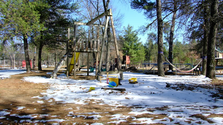 Empty back yard playground covered in thin layer of melting snow. Playground includes swing set, sandbox, wooden tower with slide and hammock next to trampoline and tree house in the background.