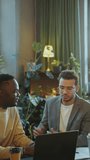 Young African American man listening to male colleague telling about business presentation on laptop when sitting together at desk in coworking office. Vertical clip
