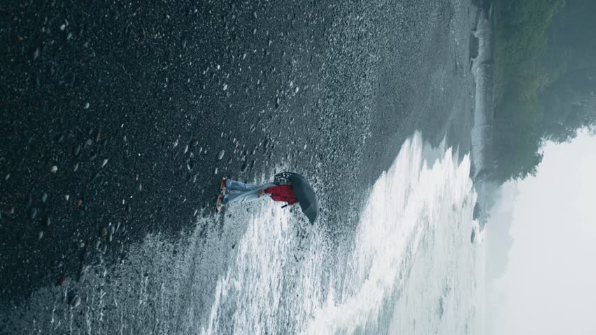 Captivating vertical video woman standing under umbrella as powerful waves crash over pebbly beach, creating mesmerizing, dynamic interaction with nature