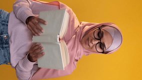 Vertical video. Learning student. Exam study. Intelligent happy smart woman in hijab glasses enjoying reading book isolated on yellow copy space background.