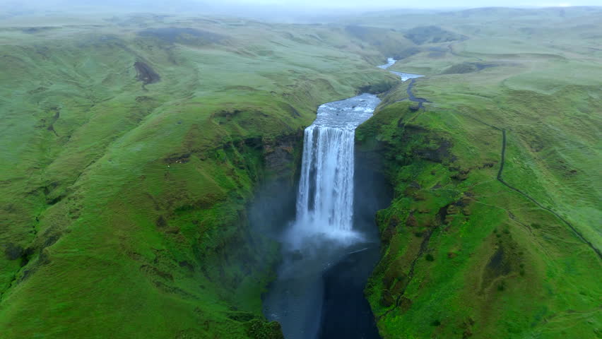 Majestic Aerial view reveals the grand scale of a powerful Skogafoss waterfall flowing through a vast green landscape, with its waters cutting a dramatic path through the terrain. Iceland, Skogar. 