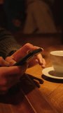 A man in a bar at a table with a cup of coffee writes SMS in a smartphone. Vertical video.