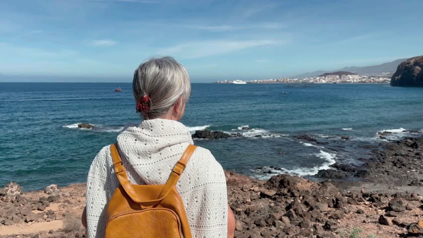 Rear view of mature grey haired woman at sea in a sunny day looking the rocky coast and the waves crashing on the beach. Senior woman in retirement or vacation enjoying freedom and nature