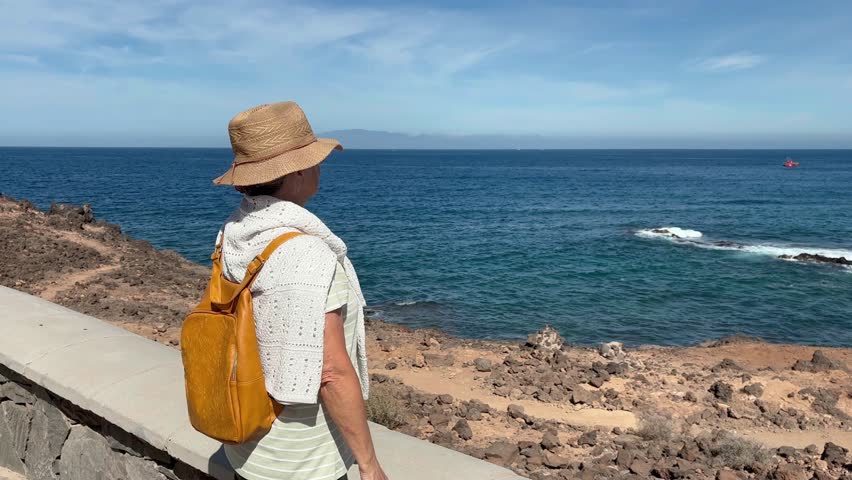 Elderly grey haired woman at sea in a sunny day looking the rocky coast and the waves crashing on the beach. Senior woman in retirement or vacation enjoying freedom and nature