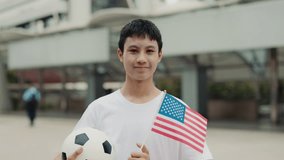 Handsome Boy Fan Standing Outside with Soccer Ball and Holding USA Flag in Hand. Asian Guy Standing and Posing for Camera with United States Flag and Holding Ball. Football and People Concept - Powered by Shutterstock - Get 15% off with code: PIKWIZARD15