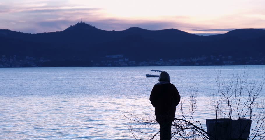 A beautiful sunset by the ocean. Small fishing boats with fisherman can be seen. An old lady is walking by the ocean.