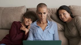 Lesbian couple and a non-binary kid using a laptop sitting on the sofa