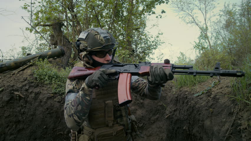 Male soldier or military man in uniform and tactical equipment sneaks up in trench with another warrior and shoots from rifle gun. Assault on enemy positions. War or military conflict. Slow motion.