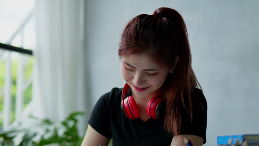 Young woman student listen music in headphone and writing on note book about lists works things to do homework