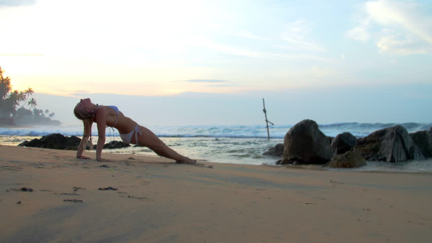 Woman in bikini improves endurance performing reverse plank pose on beach at sunset. Lady athlete enjoys yoga session against ocean