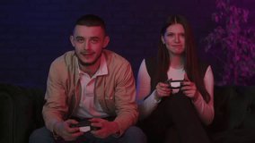 Funny young couple sitting on sofa at home and playing video games on console. Esport fans enjoying their playstation. Boyfriend don't give up and interferes his girlfriend win the game