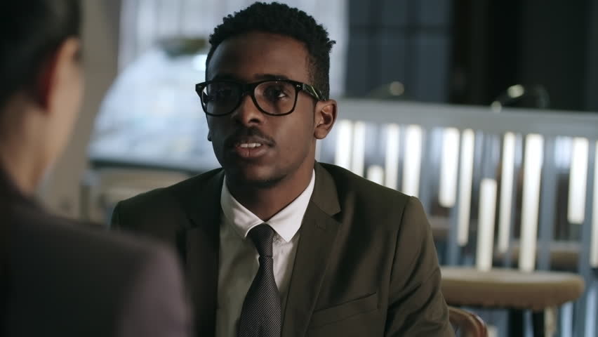 Young African businessman in eyeglasses laughing and smiling while chatting with female colleague in cafe