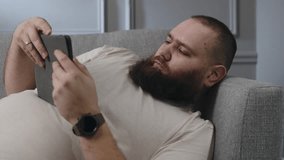 Young caucasian bearded guy watching some content on tablet in living room. Chill time at home alone. Adult man sitting couch and using tablet. Relaxed male laying down couch, watching movie