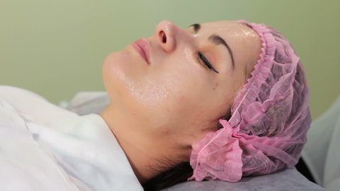 Cosmetologist applies cream on face of lying woman. Girl in bonnet lying on the session at the beautician. Woman receiving spa treatment