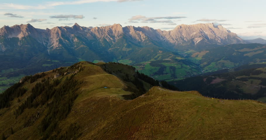 A drone flies above a green mountain slope in Austria, offering a stunning view of the majestic mountain range and the vast landscape under a clear sky