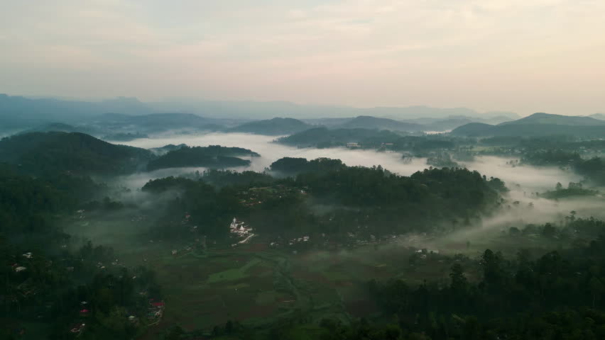 Aerial drone sweeps over misty Haputale mountains in Sri Lanka at dawn. Clouds envelop green highlands, terraced tea plantations. Sunrise illuminates serene landscape, inviting exploration, adventure.