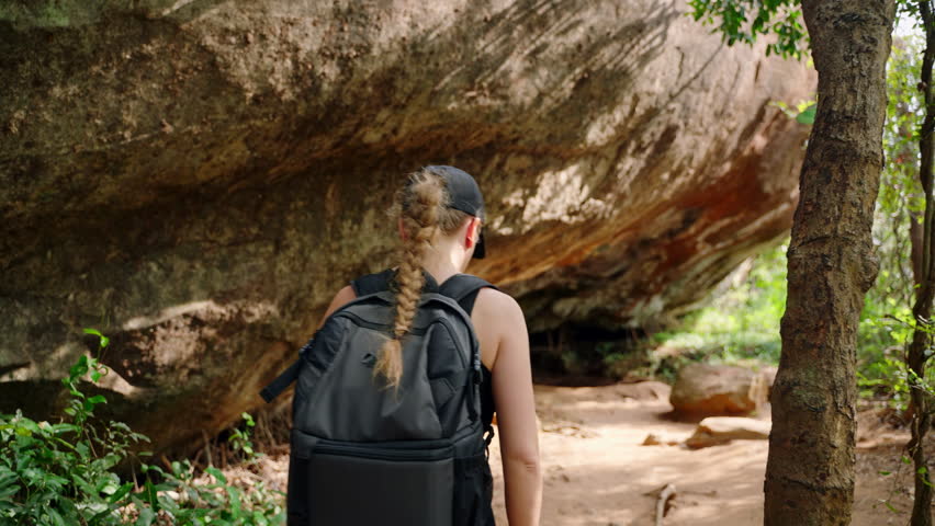 Active female hiker explores scenic grotto trail in rugged terrain. Wearing casual outdoor gear, treks beneath large rocky overhang surrounded by plants. Solo travel adventure, wilderness hike. Slowmo