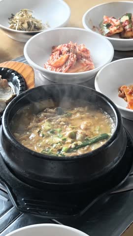 Boiling soy bean paste soup, a dish in Korean food together with other side dishes on table in a restaurant. High angle view. Vertical format. Royalty-Free Stock Footage #3498921725