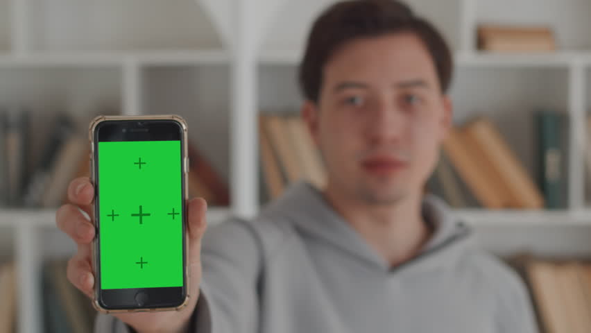 Young man in casual clothes showing a smartphone with a mockup green screen close up with the background of white bookcases with many books on the bookshelves
