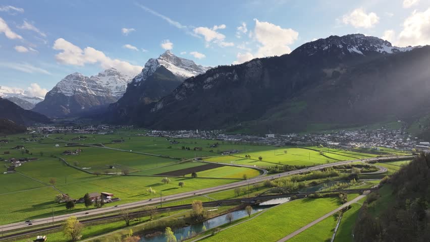 Aerial of Lush green valley near Walensee, Weesen, Switzerland, with sprawling fields and majestic snow-capped mountains under clear sky