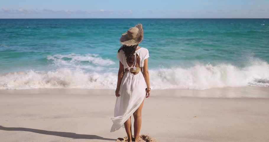 Back view of beautiful sensitive beach woman in white dress standing and looking to the sea with windy weather, a bohemian girl. High-quality travel inspirational 4k footage in slow motion