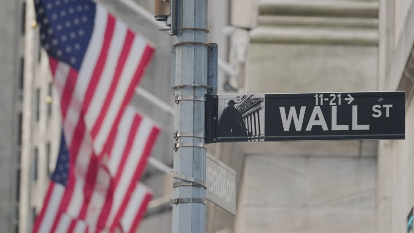 Close up of Wall Street sign in New York City with USA flags waving in background symbolizing the United States financial industry