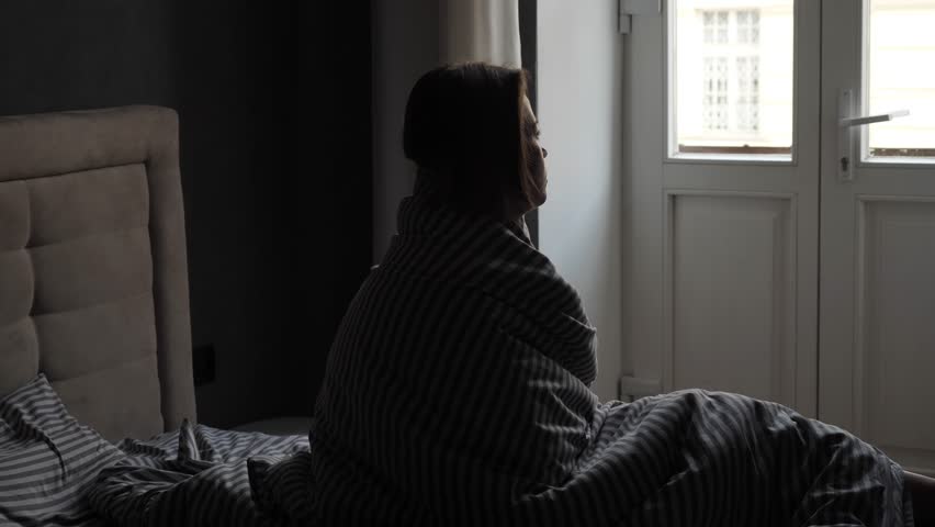 Slow motion. Silhouette of a pensive girl covered with a blanket who sits on the bed and looks out the large window. Sad or sick woman on bed in the morning. Camera movement on a gimbal.