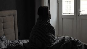 Slow motion. Silhouette of a pensive girl covered with a blanket who sits on the bed and looks out the large window. Sad or sick woman on bed in the morning. Camera movement on a gimbal. - Powered by Shutterstock - Get 15% off with code: PIKWIZARD15