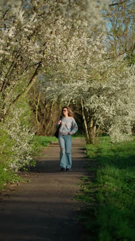 Woman enjoys spring walk among flowers. Woman walks along path in park surrounded by flowering trees. Concept of Spring, nature, pleasure.
