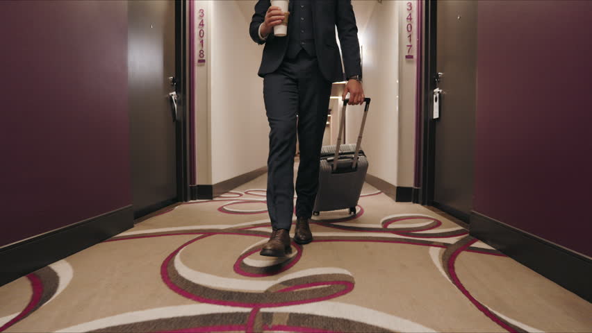 In a modern hotel corridor, a businessman walks with a coffee cup and a rolling suitcase, his stylish suit complementing the sophisticated surroundings. His steps are precise and unhurried. 