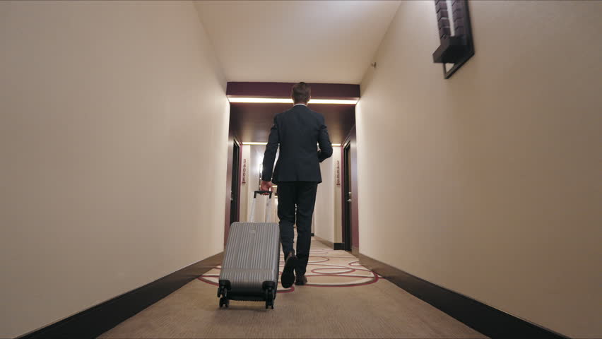 Businessman in a sleek, dark suit walks down a hotel corridor, coffee in one hand and a suitcase in another. His brisk, determined steps showcase his mastery of balancing travel and tasks. 