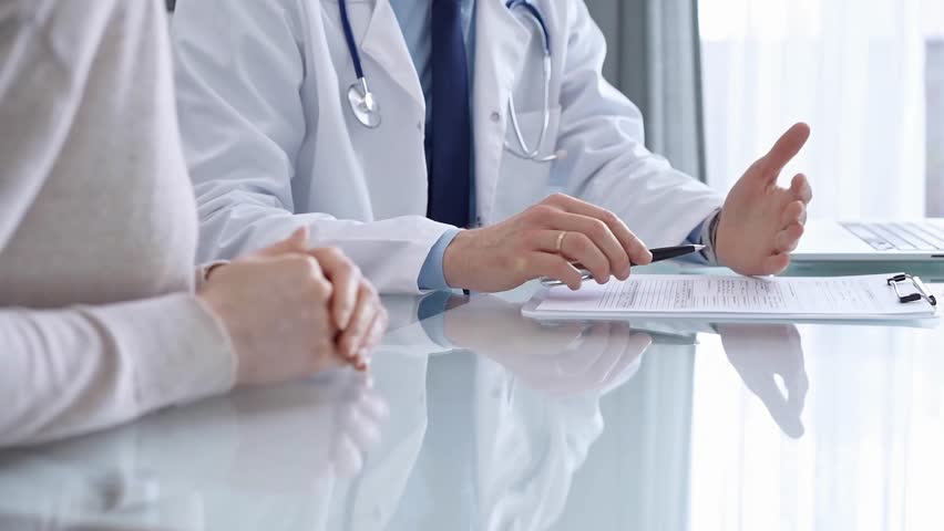 Doctor is consulting patient in modern clinic. Healthcare professional discusses medical information with a woman, gesturing above the glass desk. Medicine