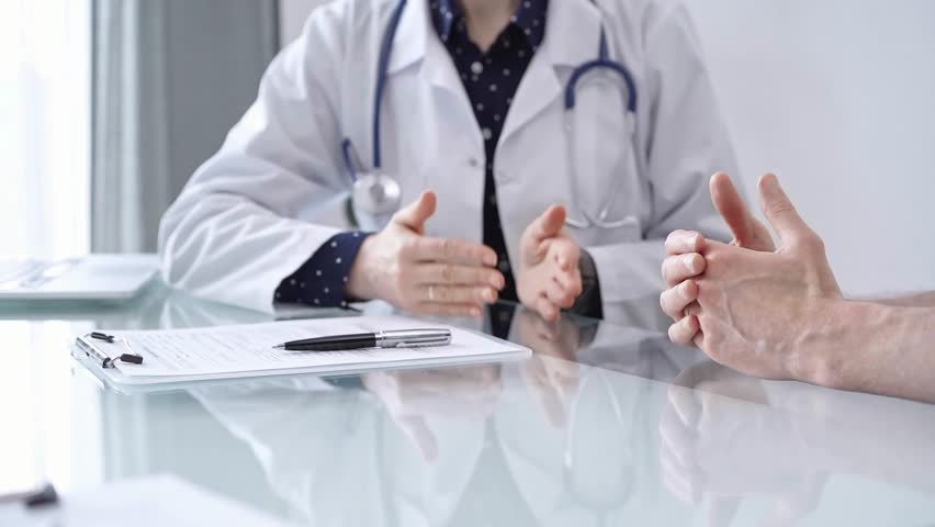 Doctor is consulting patient in modern fair clinic. Healthcare professional discusses medical information with a man, gesturing above the glass desk. Medicine