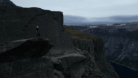 Capture breathtaking views of Trolltunga, Norway, as a lone figure stands atop the iconic rock formation, framed by majestic fjords, in this stunning aerial footage.