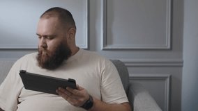 Adult young bearded man using portable tablet and lying down sofa. Male sitting couch and browsing internet pages with tablet. Addicted guy watching favorite movie with chips. Can't stop eating crisps