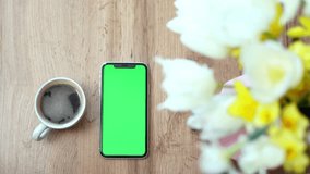 Close-up of a smartphone with green screen near cup with coffee on flowers background. Press browsing chroma key online, typing text, reading social media. 4k footage