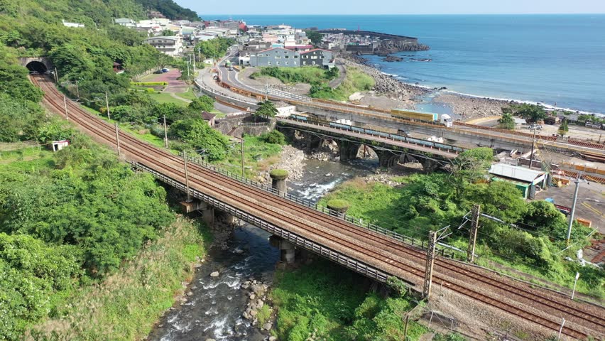 Aerial view of a Puyuma Express train dashing across a river by the mountainside and a coastal highway running through a seaside village, in Toucheng Township, Yilan County, Taiwan