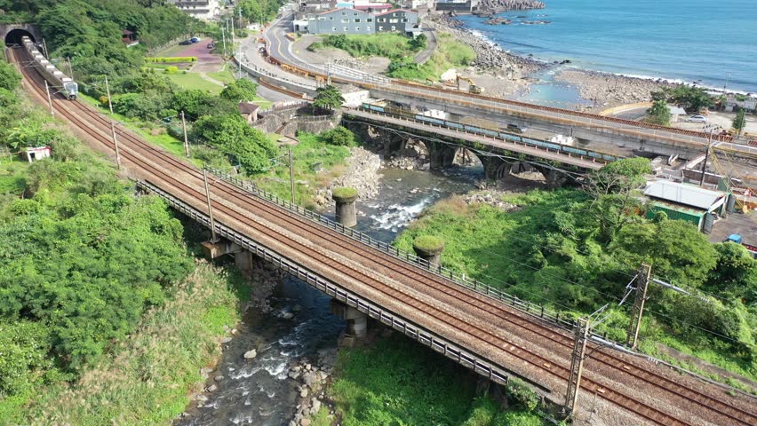 Aerial view of a fast EMU900 train dashing across a river by the mountainside and a coastal highway running thru a seaside village next to Daxi Harbor on a sunny summer day, in Toucheng, Yilan, Taiwan