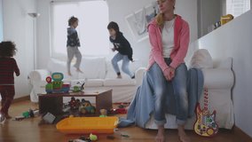 Exhausted mother sits with eyes closed and lying back on a couch, surrounded by her playful children and a disheveled living room strewn with toys, slow motion. - Powered by Shutterstock - Get 15% off with code: PIKWIZARD15