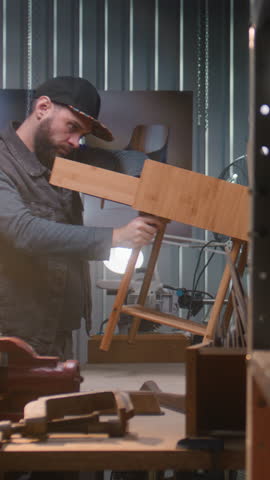 Male carpenter makes or assembles wooden bedside table. Young craftsman, artisan works in modern furniture workshop. Board with tools and equipment on the wall. Handmade and entrepreneurship concept.