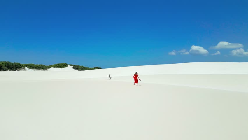Aerial view of Lady in red dress spin around on white sand dunes in Brazil desert in Parque Nacional dos Lencois Maranhenses. Attractive young woman in red dress.